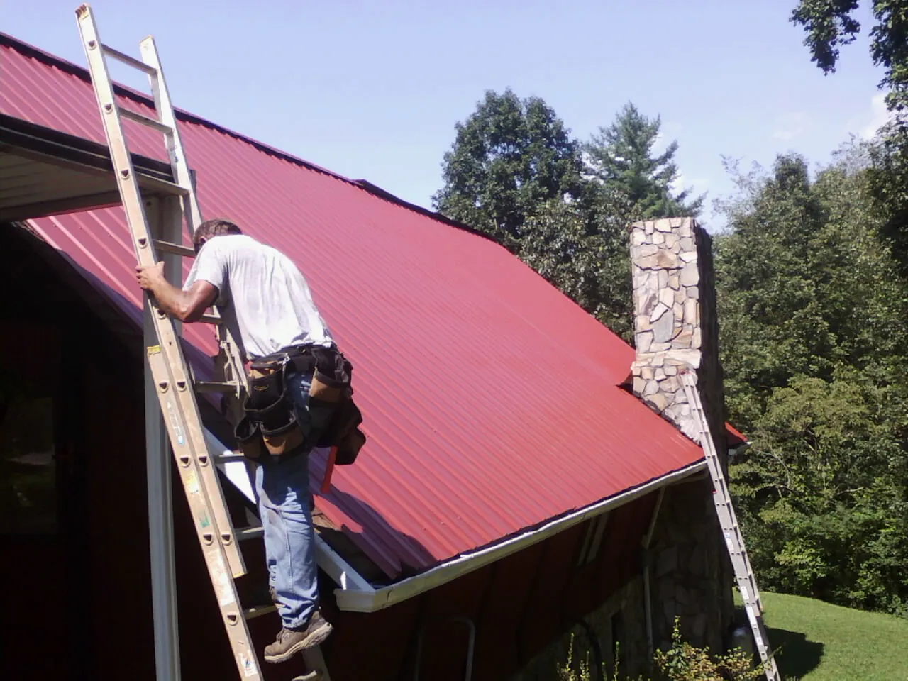 Roofer on a ladder beside a red metal roof and stone chimney — Soffit & Fascia Repair by Roofing Brothers in Rimersburg